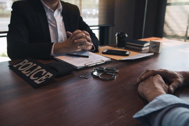A detective and a man are talking, with handcuffs on the table between them