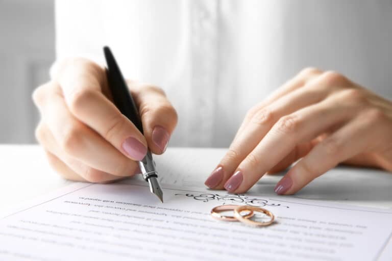 Close-up of a woman signing a legal document with two wedding rings placed on the paper.