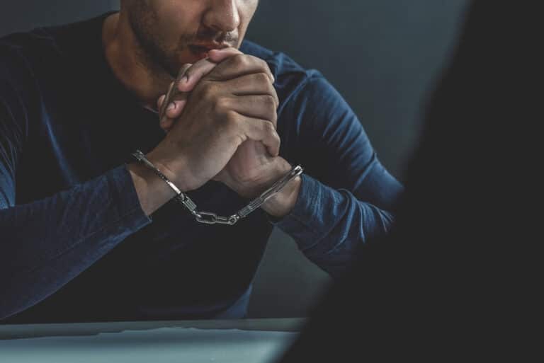 A man with handcuffs in an interrogation room being interviewed.